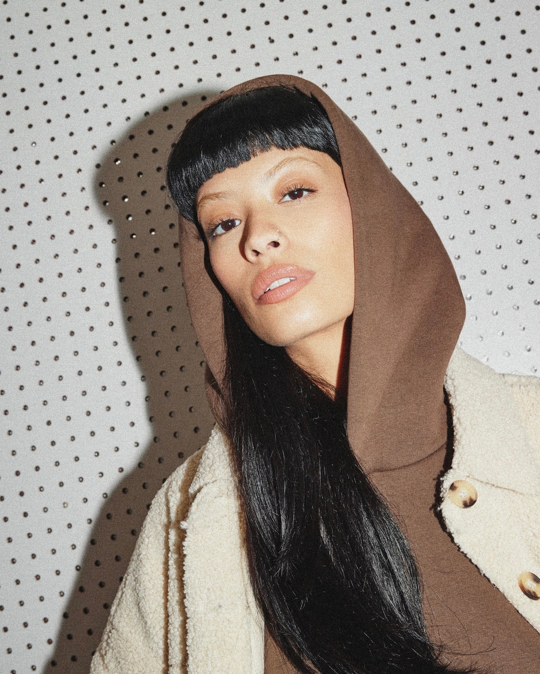 Woman with a fringe hairstyle poses in a brown hooded sweatshirt and off-white button-down jacket against a dotted backdrop.