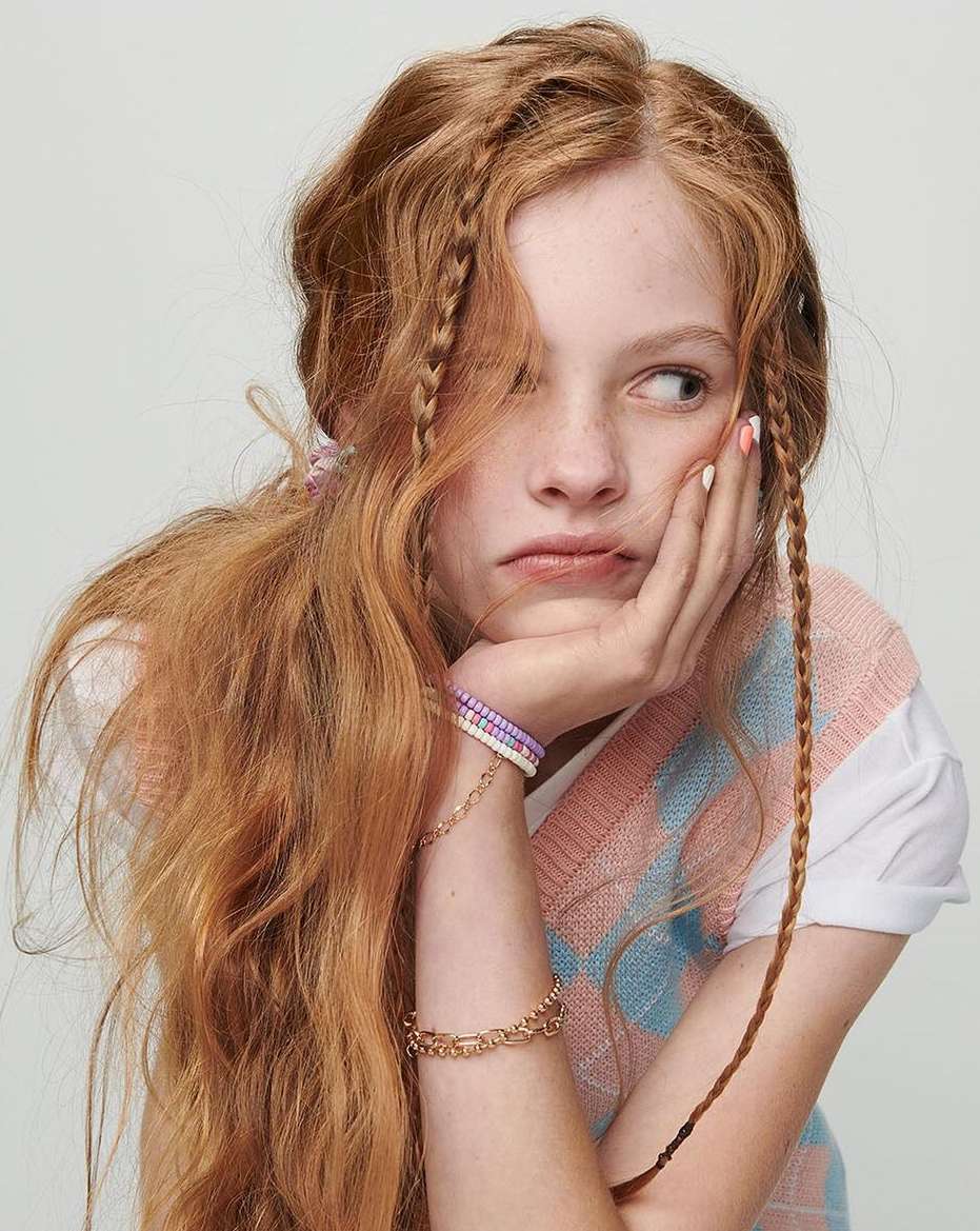 Portrait of a young woman with long red hair, light skin, and pouting expression, wearing a sweater vest with bracelets, against a grey background.