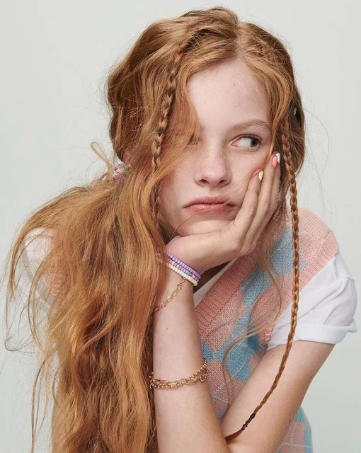 Portrait of a young woman with long red hair, light skin, and pouting expression, wearing a sweater vest with bracelets, against a grey background.