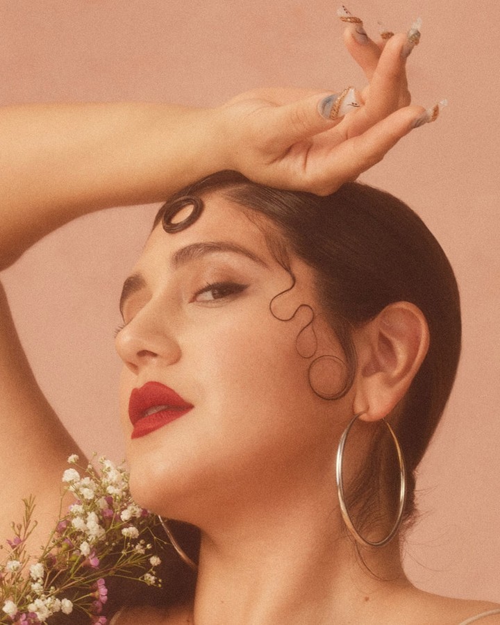 Close-up portrait of a woman with red lipstick, sculptural baby hair, hoop earrings, and hand raised to her forehead against a muted pink background.