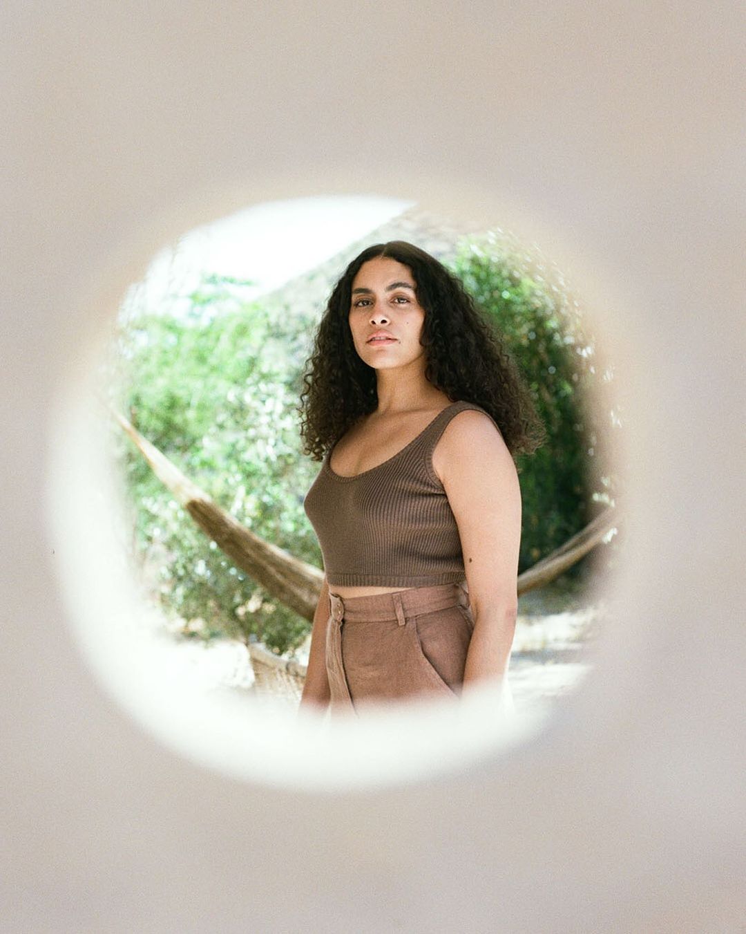 Woman with curly hair poses in brown crop top and pants, framed by white circular wall opening, with outdoor greenery in the background.