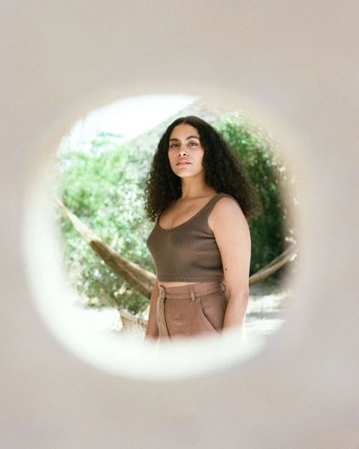 Woman with curly hair poses in brown crop top and pants, framed by white circular wall opening, with outdoor greenery in the background.