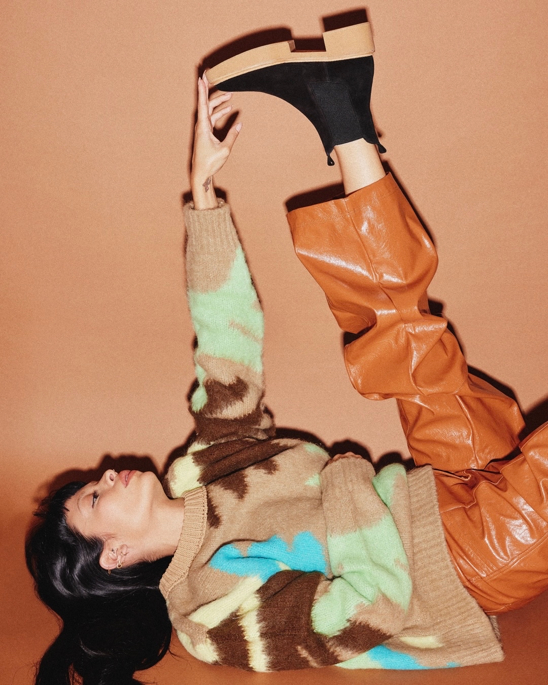 A woman in a patterned sweater and brown faux-leather pants reclines against an orange background, holding up a black platform shoe.