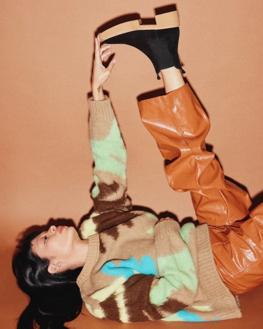 A woman in a patterned sweater and brown faux-leather pants reclines against an orange background, holding up a black platform shoe.