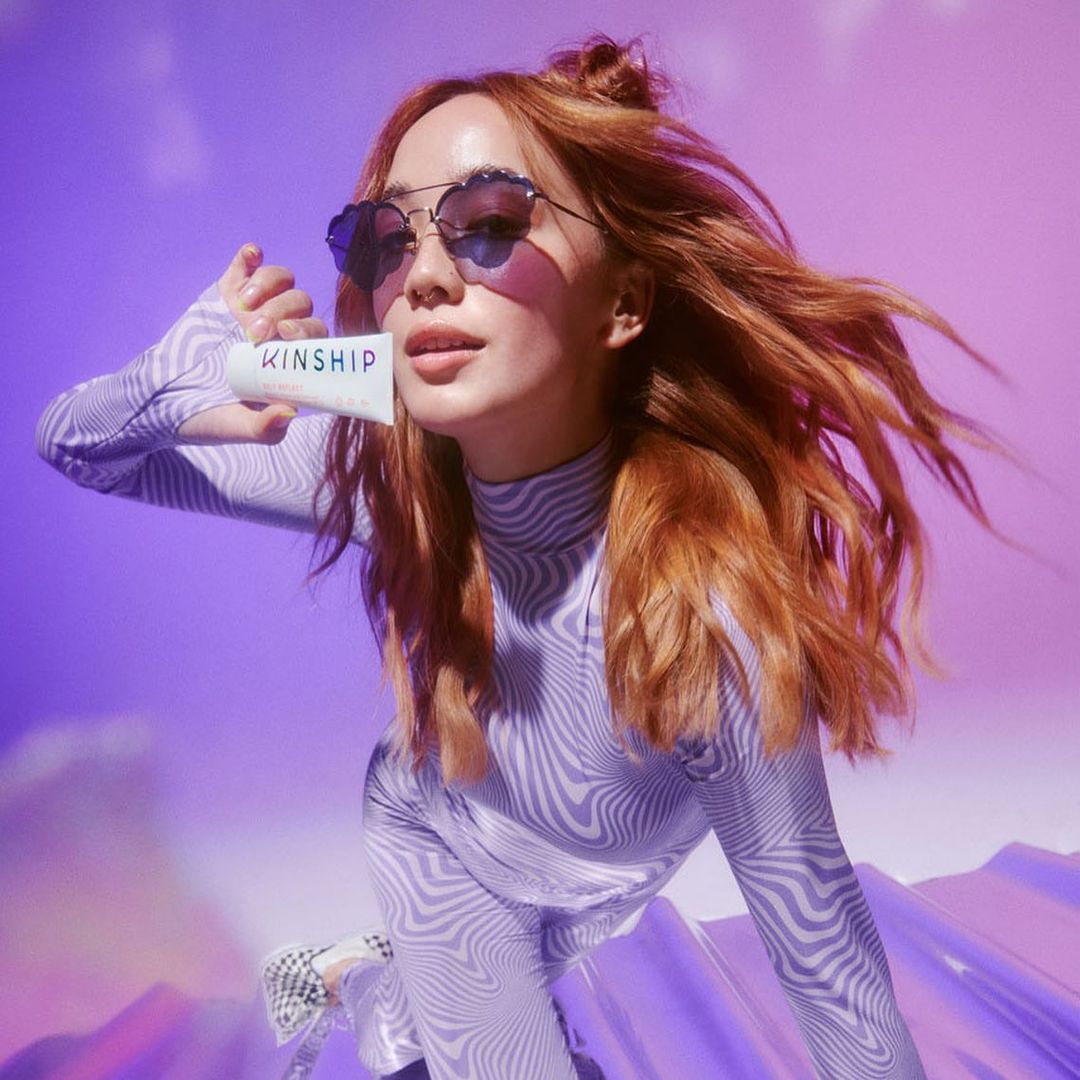 Woman with red hair in patterned top and heart-shaped sunglasses holds a white Kinship product tube against a purple background.