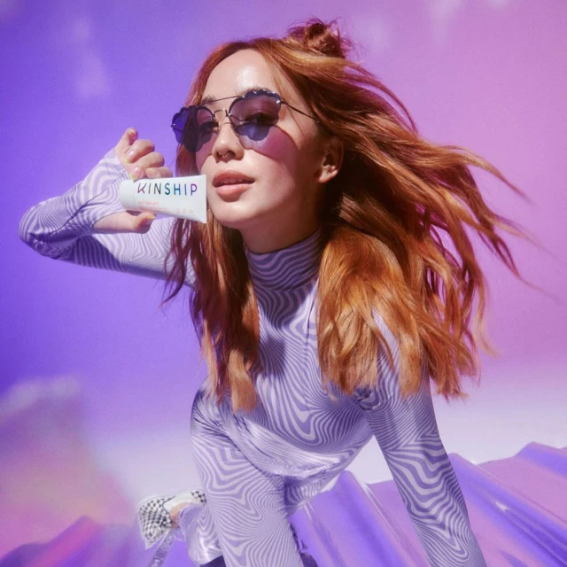 Woman with red hair in patterned top and heart-shaped sunglasses holds a white Kinship product tube against a purple background.