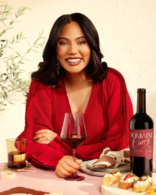 Smiling woman in a red dress poses indoors with a glass of red wine and a bottle of Domaine Curry Cabernet Sauvignon on a table.