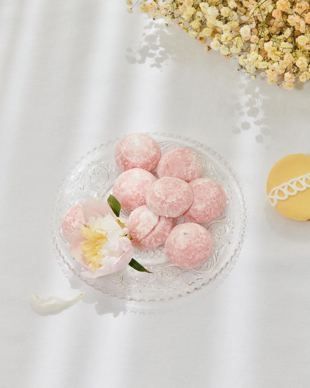 Overhead shot of a clear plate of pink mochi and a flower, accented by smaller blossoms and a yellow confectionary on a white surface.