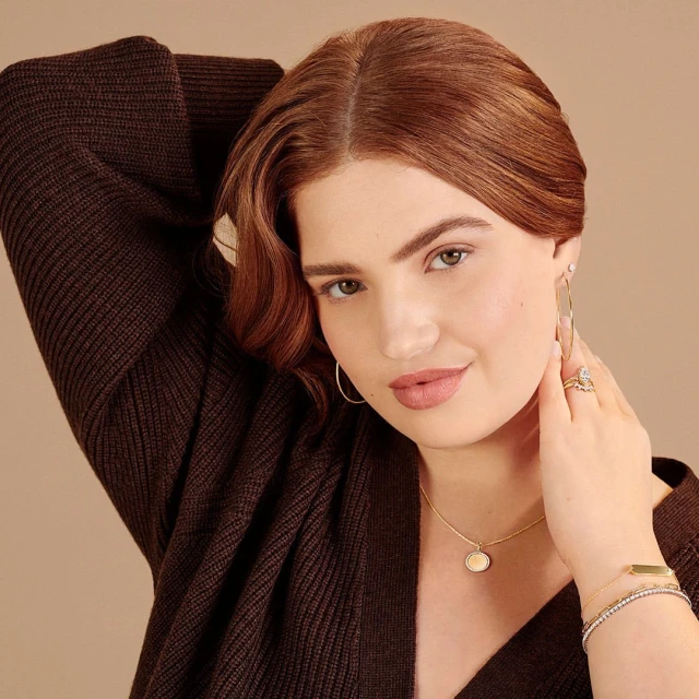 Attractive red-haired woman in dark brown cardigan and hoop earrings, wearing a gold necklace and bracelets in a studio shot.