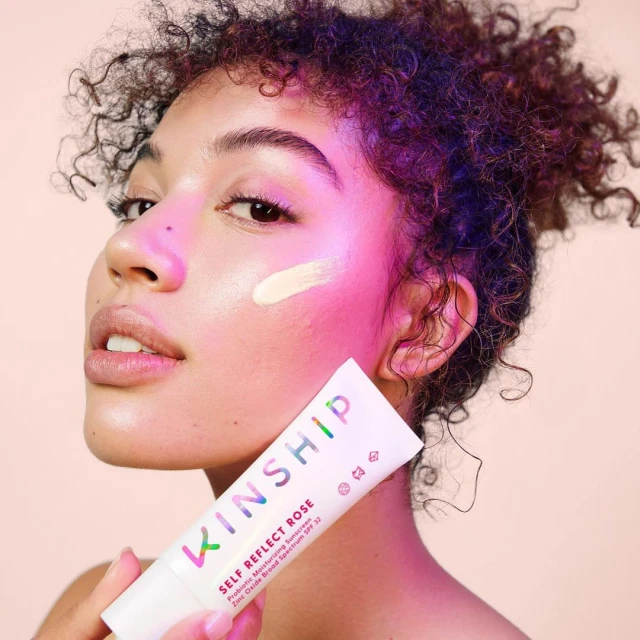 Attractive African American woman with glowing skin applying sunscreen on her face while holding tube of Kinship Self Reflect Rose SPF 32.