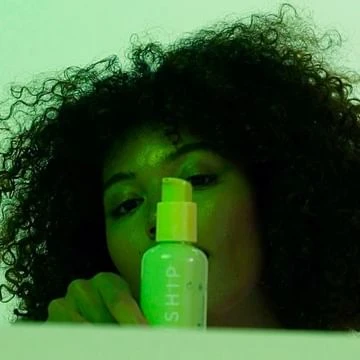 Adult female with curly hair holding a lime green bottle, partially obscured by the bottle, in an indoor setting with green lighting.