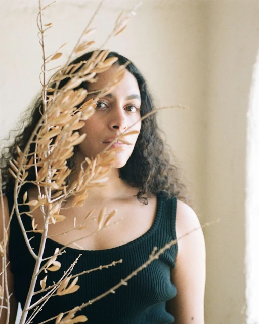 Portrait of a woman with curly hair in a black top, partially obscured by a branch with dry leaves, against a neutral indoor background.