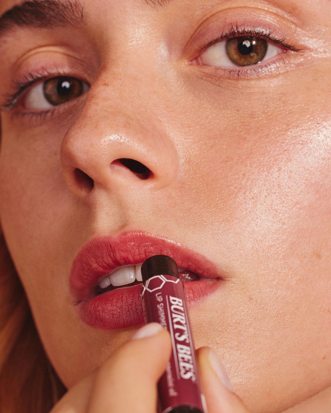 Close-up of an adult woman applying shimmering lip balm with a tube of Burt's Bees lip shimmer, highlighting the product's texture and effect.