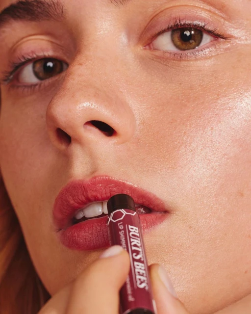 Close-up of an adult woman applying shimmering lip balm with a tube of Burt's Bees lip shimmer, highlighting the product's texture and effect.