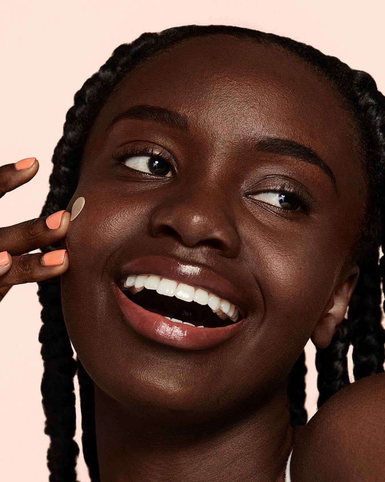 Close-up shot of a smiling Black woman with a skincare patch on her cheek, wearing her hair in braids, against a light pink backdrop.