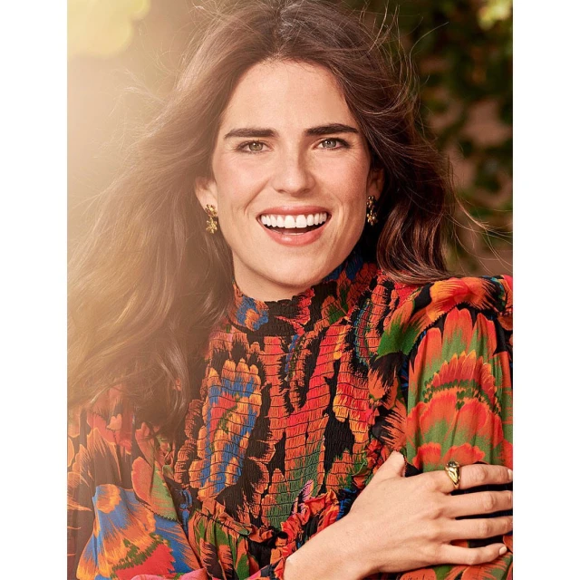 A smiling woman with long brunette hair wears a colorful floral dress and gold jewelry, posing outdoors in natural sunlight.