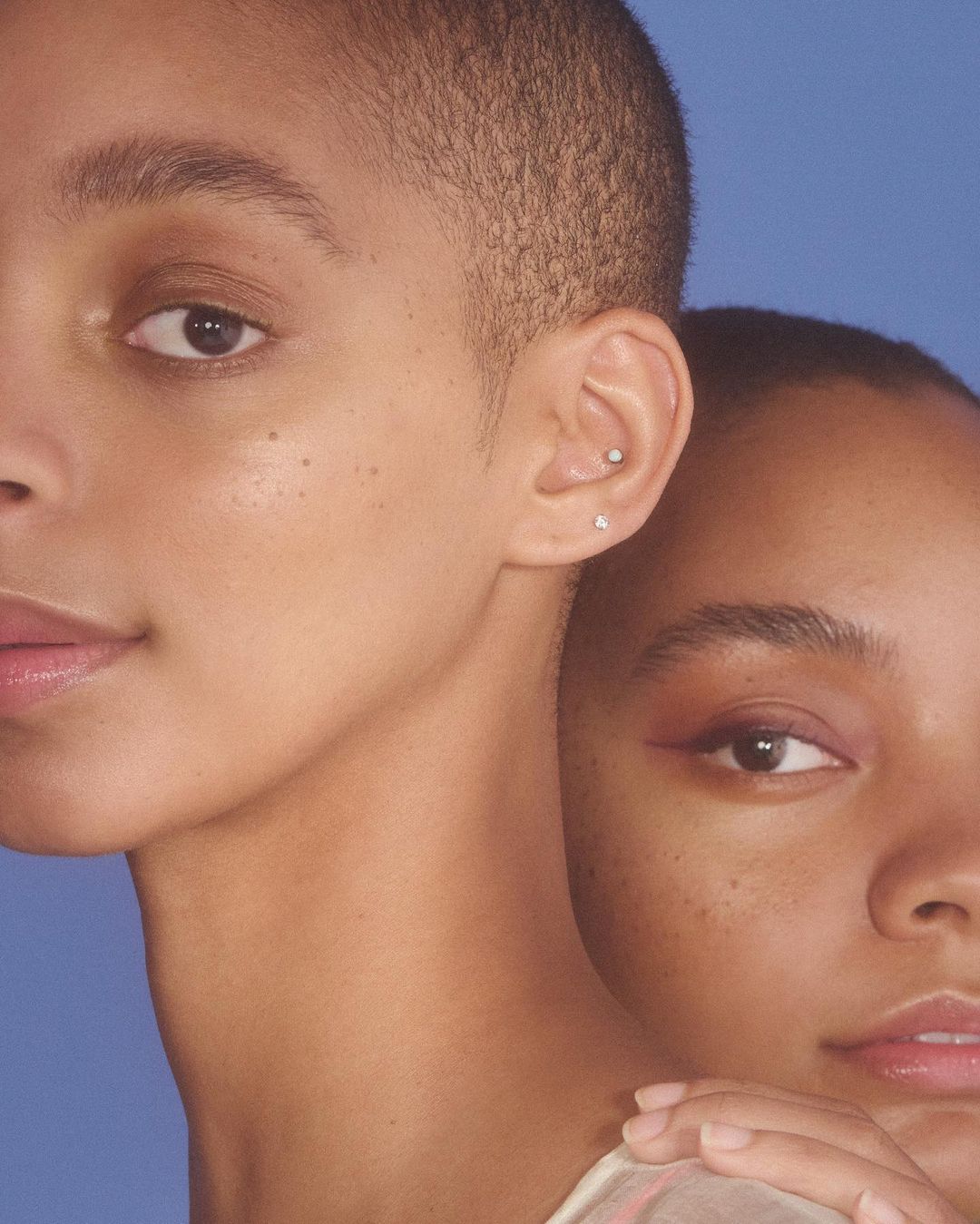 Close-up portrait of two adult women with short hair against a blue background. One woman has reddish eye makeup and freckles.