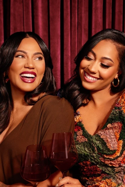 Two women smiling, holding red wine glasses against a red velvet curtain backdrop, showcasing beauty, fashion, and intimate gathering.