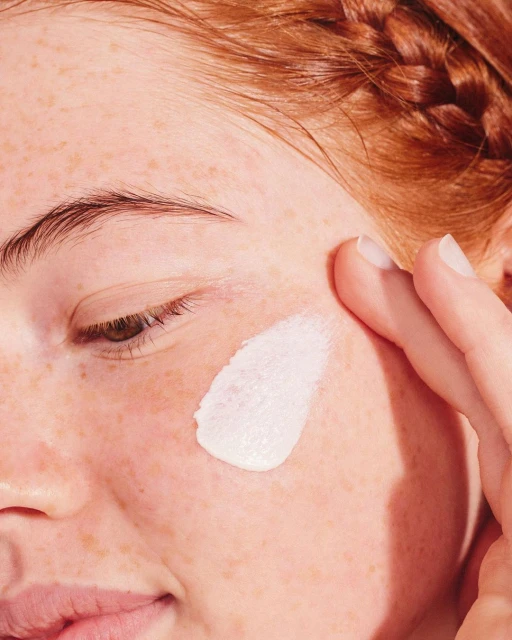 Close-up of woman with red hair applying face cream to her cheek with her fingers, showcasing skincare and beauty routine.