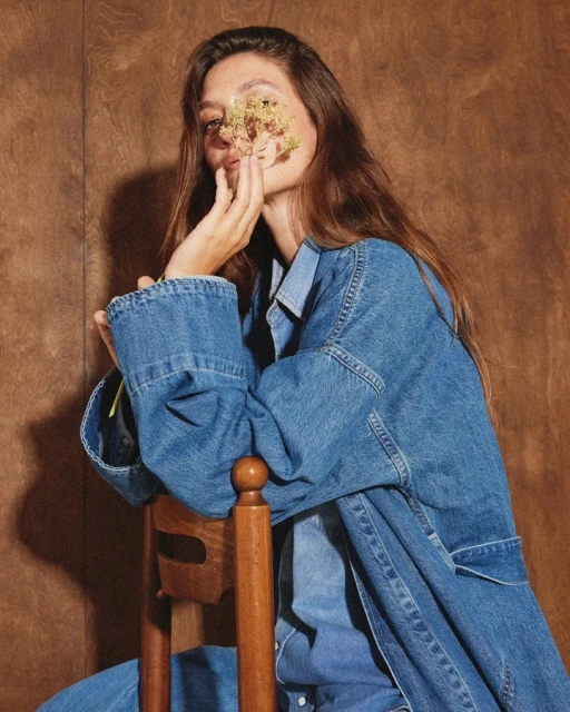 A brunette woman in a denim jacket sits on a wooden chair, holding a sprig of flowers up to her face against a wood background.