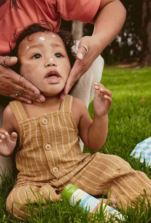 A parent applying sunscreen to the face of a toddler with dark hair, while sitting on grass outdoors in soft, natural lighting.