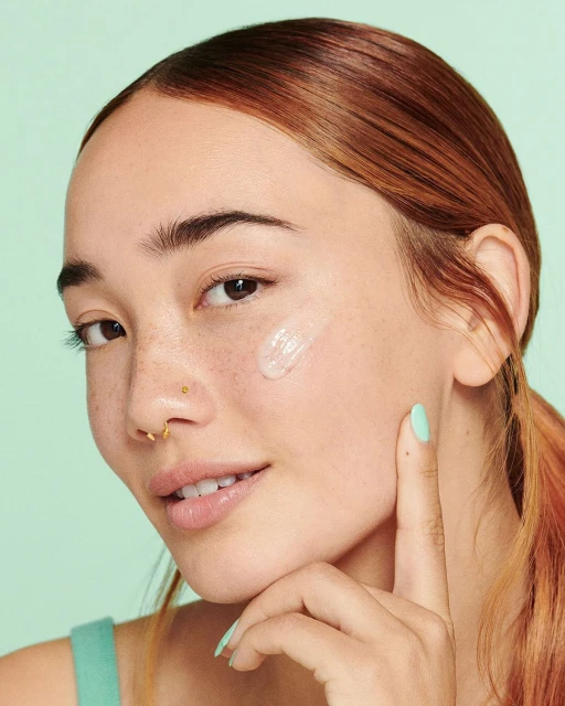 Close-up of a young woman applying a clear gel skincare product to her cheek with her finger, posing against a light-green backdrop.