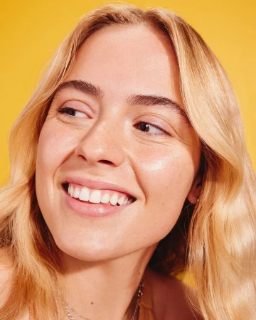 Close-up portrait of a young, smiling blonde woman with fair skin against a bright yellow background, showcasing beauty and natural light.