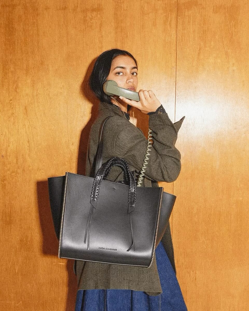 Woman in a jacket and skirt holds a telephone receiver while carrying a black leather tote bag against a wood-paneled backdrop.