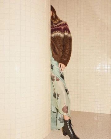 Model wearing patterned knit sweater and floral skirt partly hidden behind tiled pillar