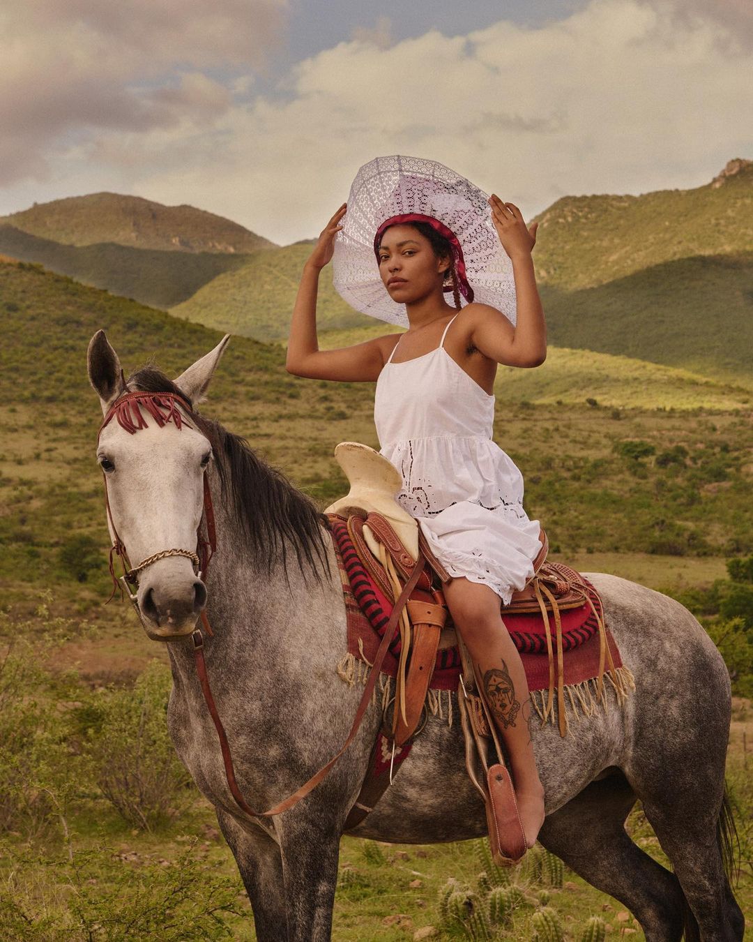 A woman in a white dress and wide-brimmed hat sits astride a gray horse in a rural setting with grassy fields and distant mountains.