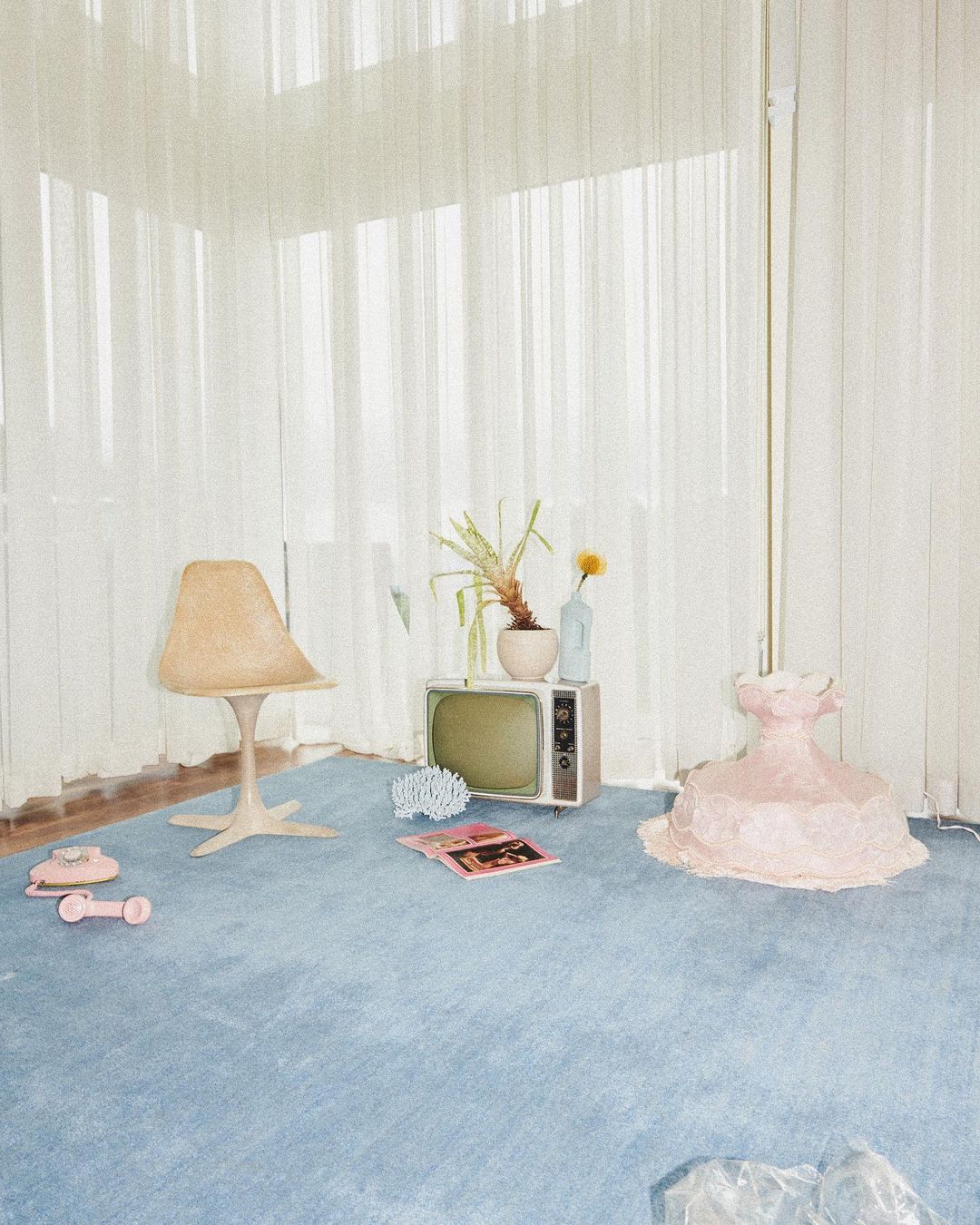 Retro-inspired interior featuring a blue carpet, vintage television, beige chair, pink sculptural form, and sheer white curtains.