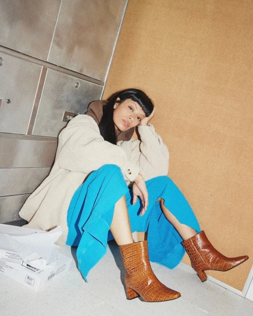 A woman sits indoors wearing an off-white jacket, brown hoodie, turquoise wide-leg pants, and brown snake-print heeled ankle boots.