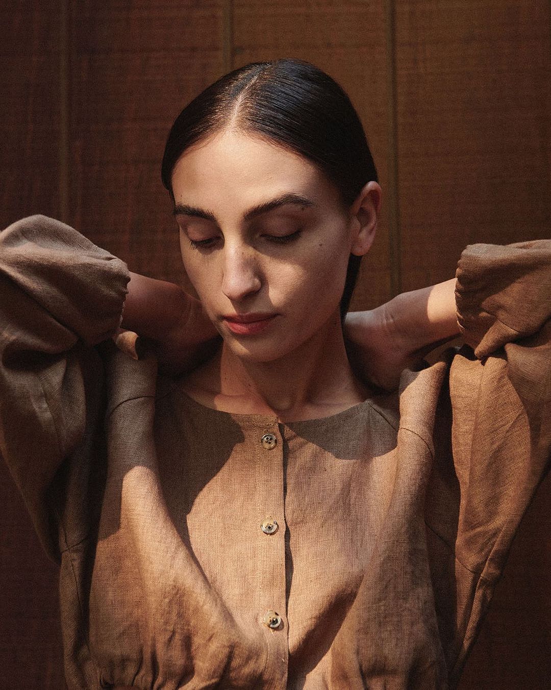 Portrait of a brunette woman with her hands behind her neck, wearing a light brown linen button-down shirt in a photo studio.