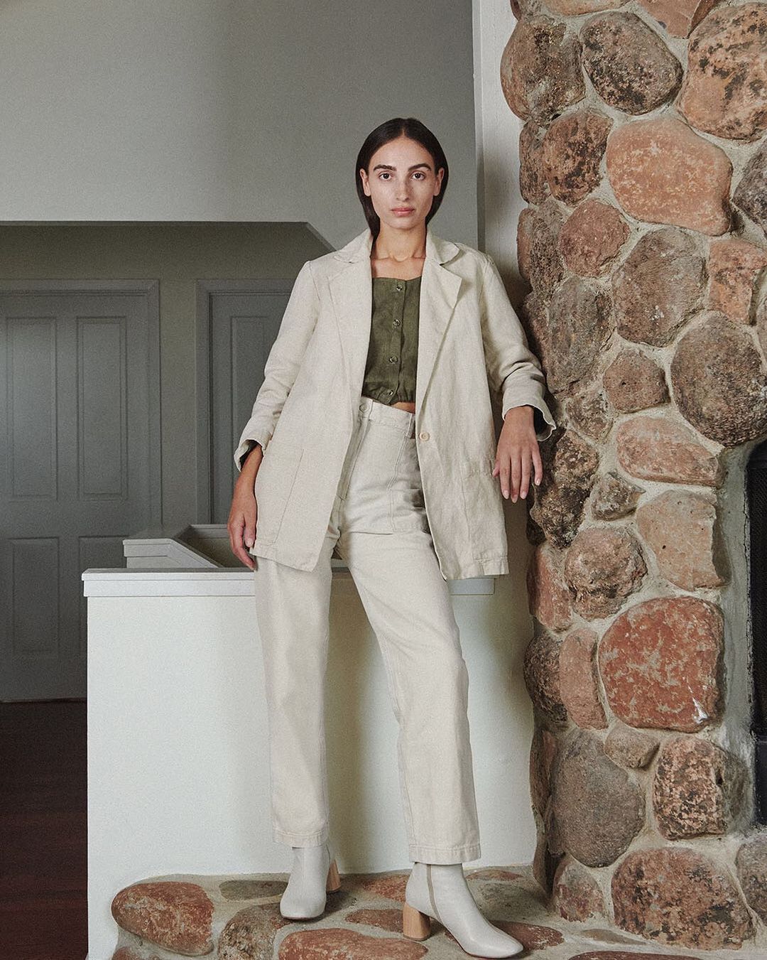 A woman posing indoors in an off-white blazer, matching pants, and heeled boots stands against a stone wall and doorway, looking directly at the camera.