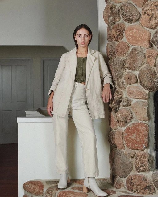 A woman posing indoors in an off-white blazer, matching pants, and heeled boots stands against a stone wall and doorway, looking directly at the camera.
