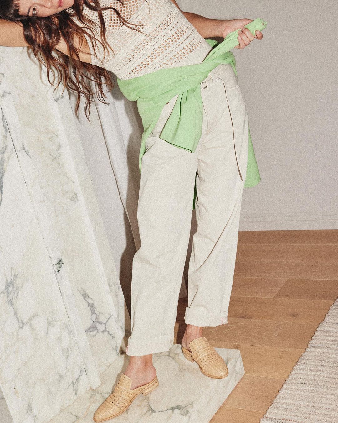 Indoor shot of a young woman wearing a knit top, casual pants, and woven mules, posing near a marble block and light wood flooring.