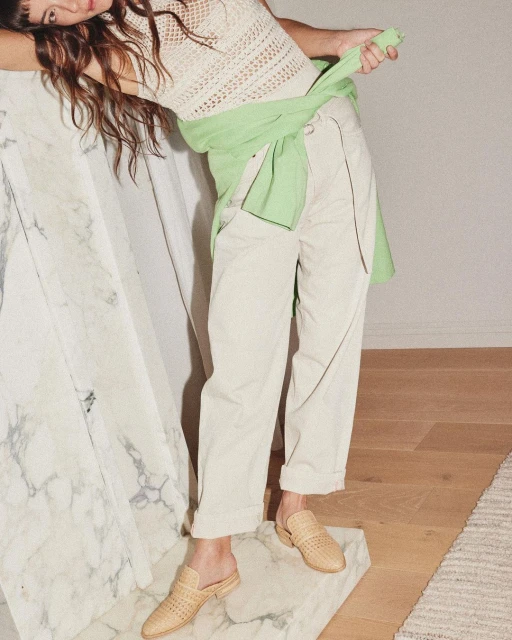 Indoor shot of a young woman wearing a knit top, casual pants, and woven mules, posing near a marble block and light wood flooring.