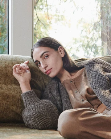 Adult woman rests on an olive-colored sofa near a window. She wears a brown sweater and pants, sitting in natural light.