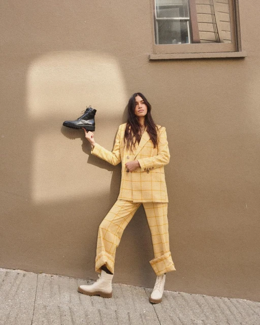 Adult female in a yellow plaid suit balances a black boot on her finger while standing outdoors against a neutral wall and concrete.