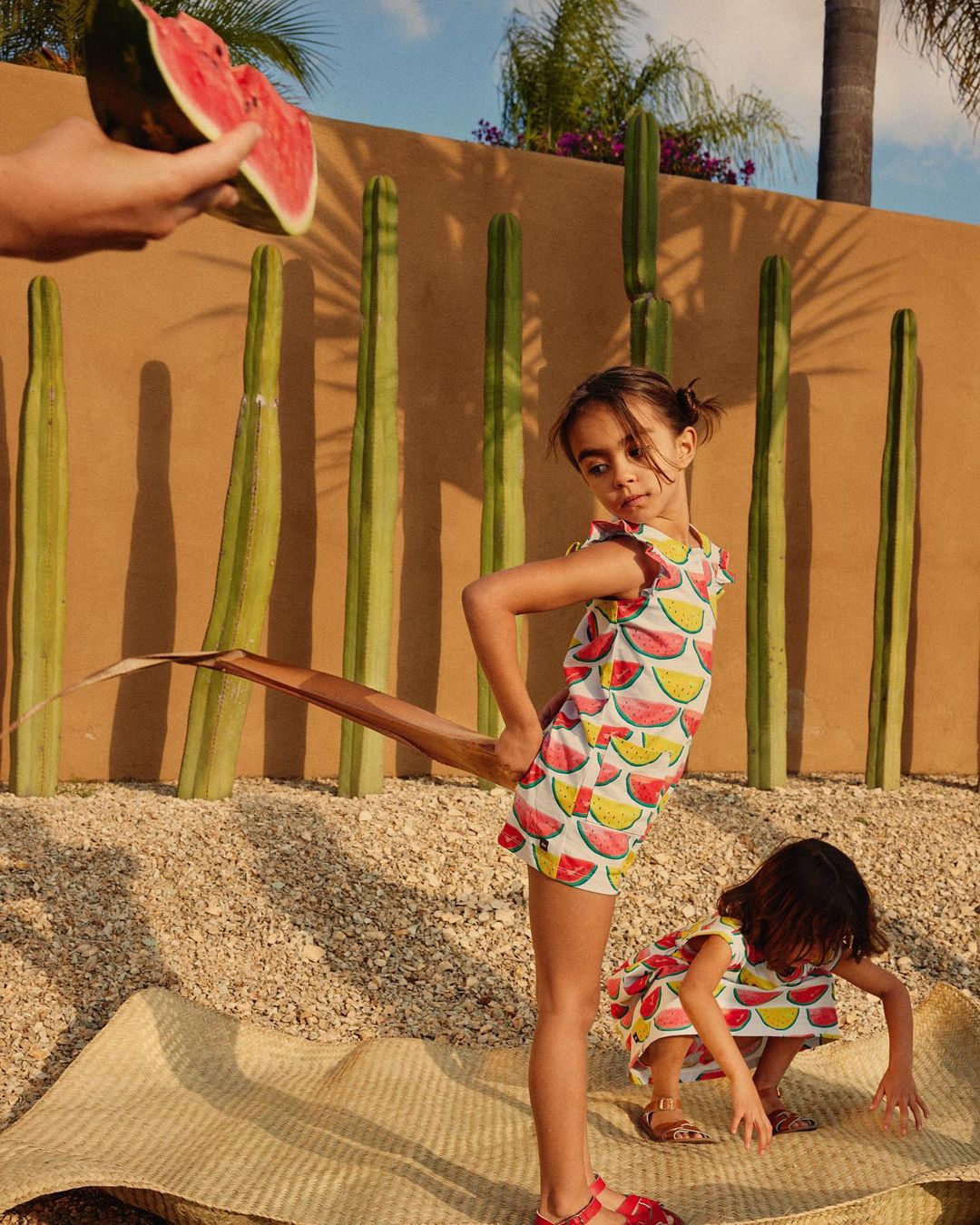 Two young girls in watermelon-print dresses pose outdoors near tall cacti and a beige wall, with a hand holding watermelon slices.