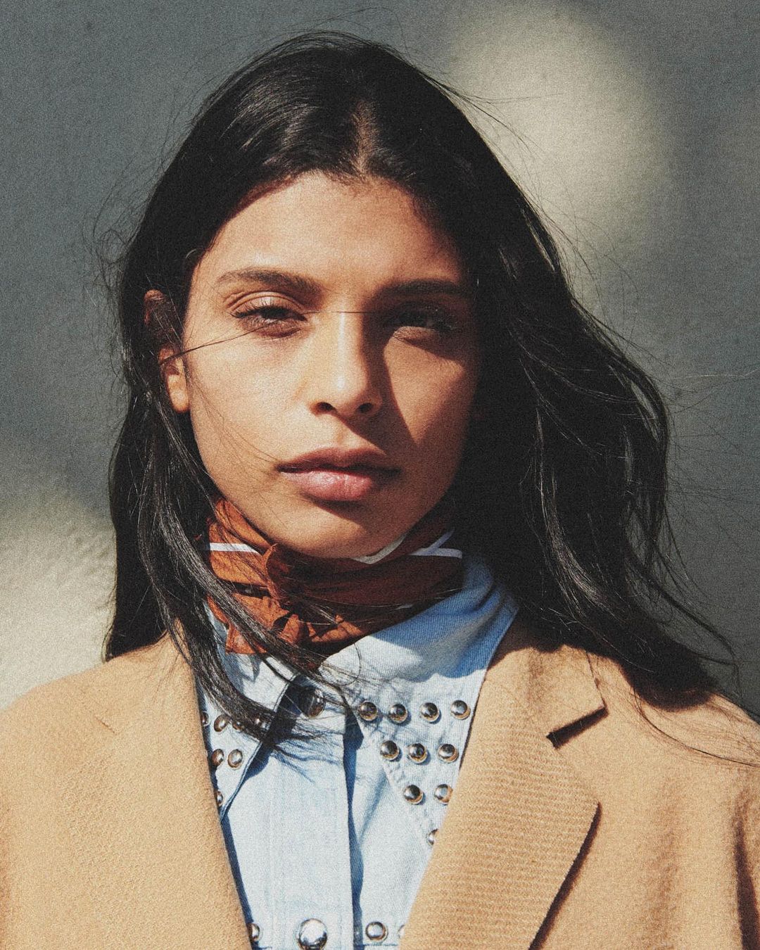 Portrait of a fashionable young adult woman with dark hair, wearing a brown jacket and light blue shirt, posing indoors with natural light.