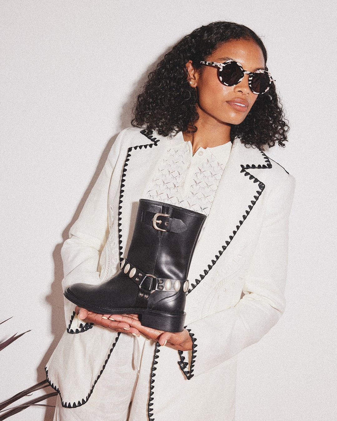 Adult woman wearing a white blazer with black trim holding a black leather boot with silver accents in a studio with a white background.