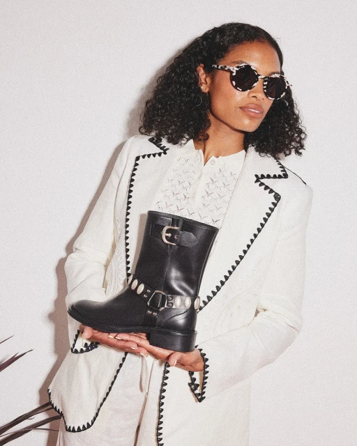 Adult woman wearing a white blazer with black trim holding a black leather boot with silver accents in a studio with a white background.