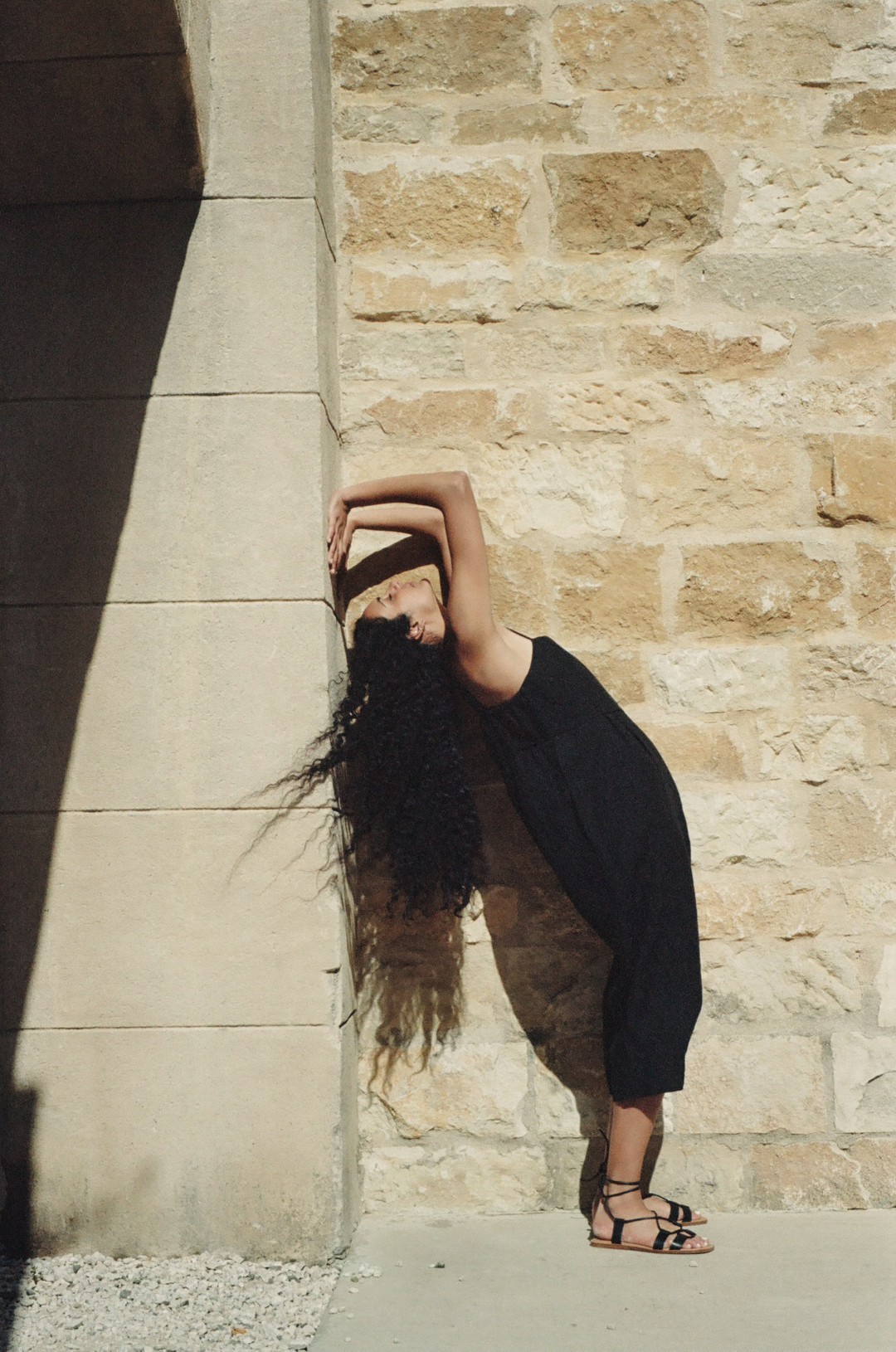 A woman in a black jumpsuit poses against a tan stone wall with dark shadows. She has long, curly hair blowing to the side.