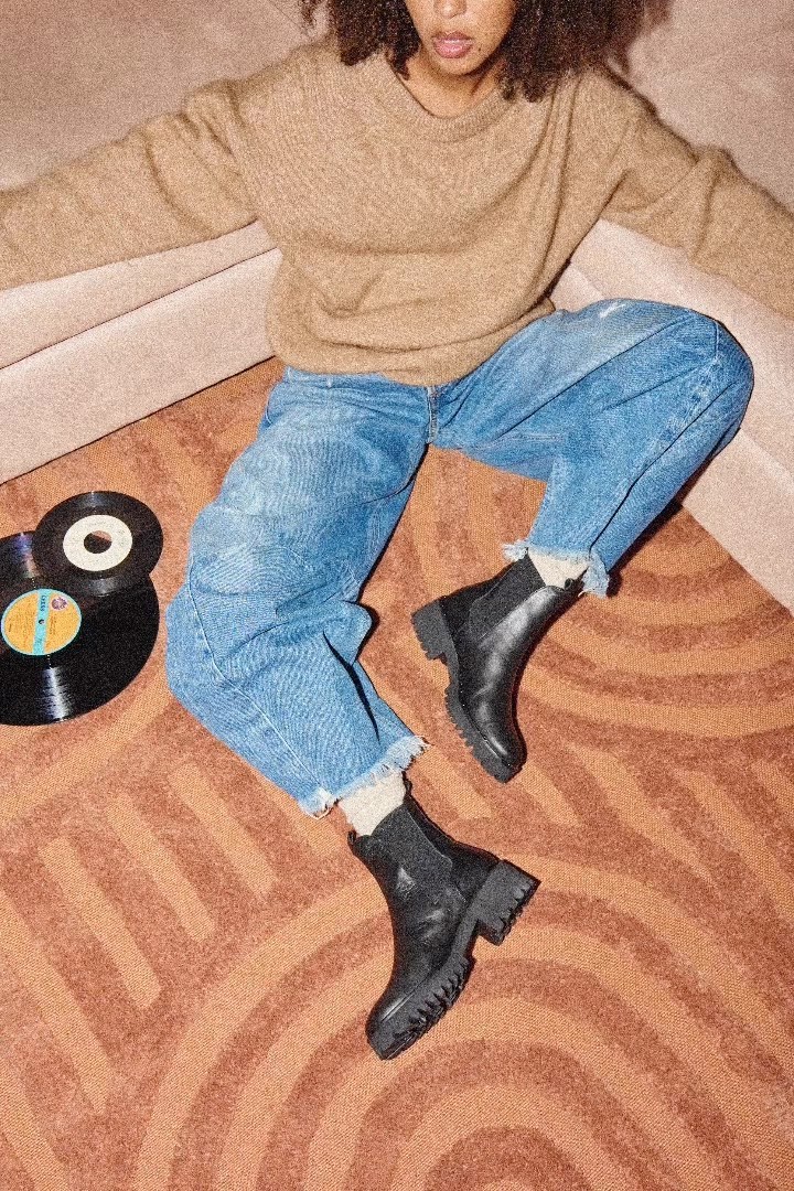 Fashion shot of a woman wearing a sweater, jeans, and boots sitting on a carpet with vinyl records in a neutral indoor setting.