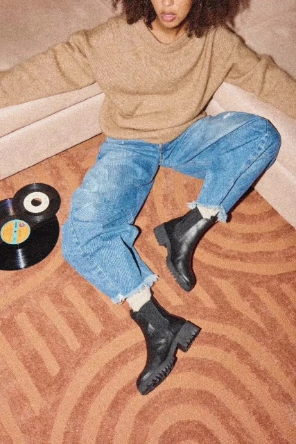 Fashion shot of a woman wearing a sweater, jeans, and boots sitting on a carpet with vinyl records in a neutral indoor setting.