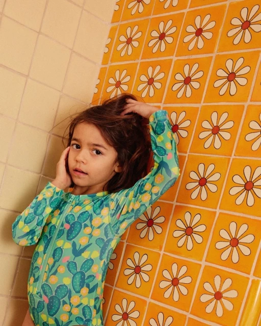 Young child in patterned swimsuit stands in front of daisy-patterned tile wall, reaching for her hair in a brightly lit indoor space.