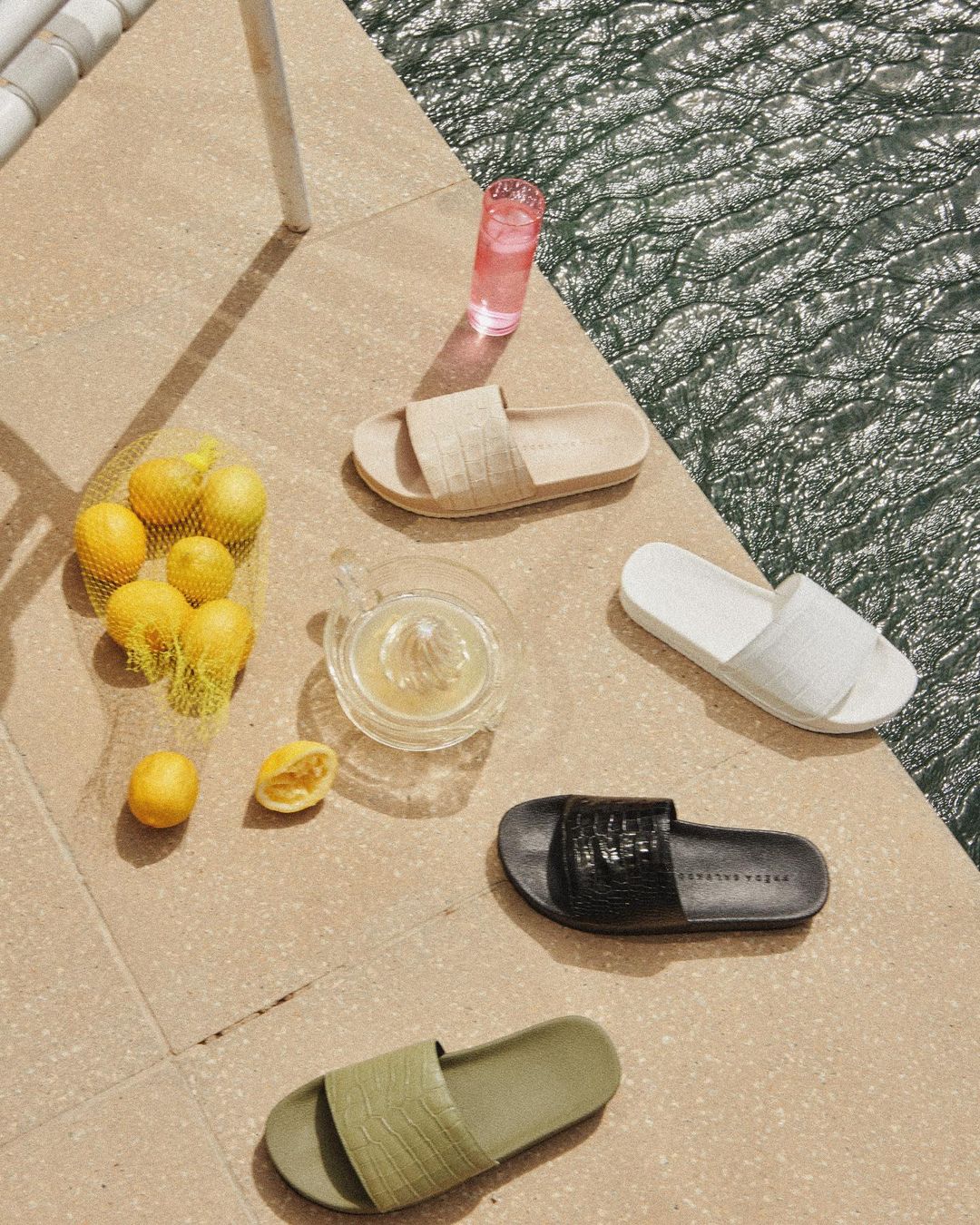 Still life of slide sandals on a tile surface with a glass of pink liquid, lemons in a net bag, and a citrus press.
