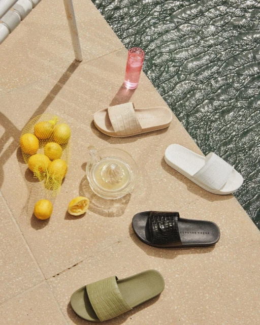 Still life of slide sandals on a tile surface with a glass of pink liquid, lemons in a net bag, and a citrus press.
