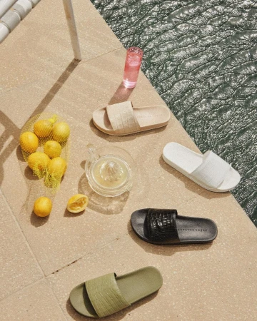 Still life of slide sandals on a tile surface with a glass of pink liquid, lemons in a net bag, and a citrus press.